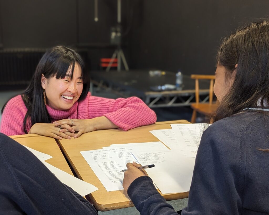 Sophie (in pink) crouching at a desk and laughing with a student