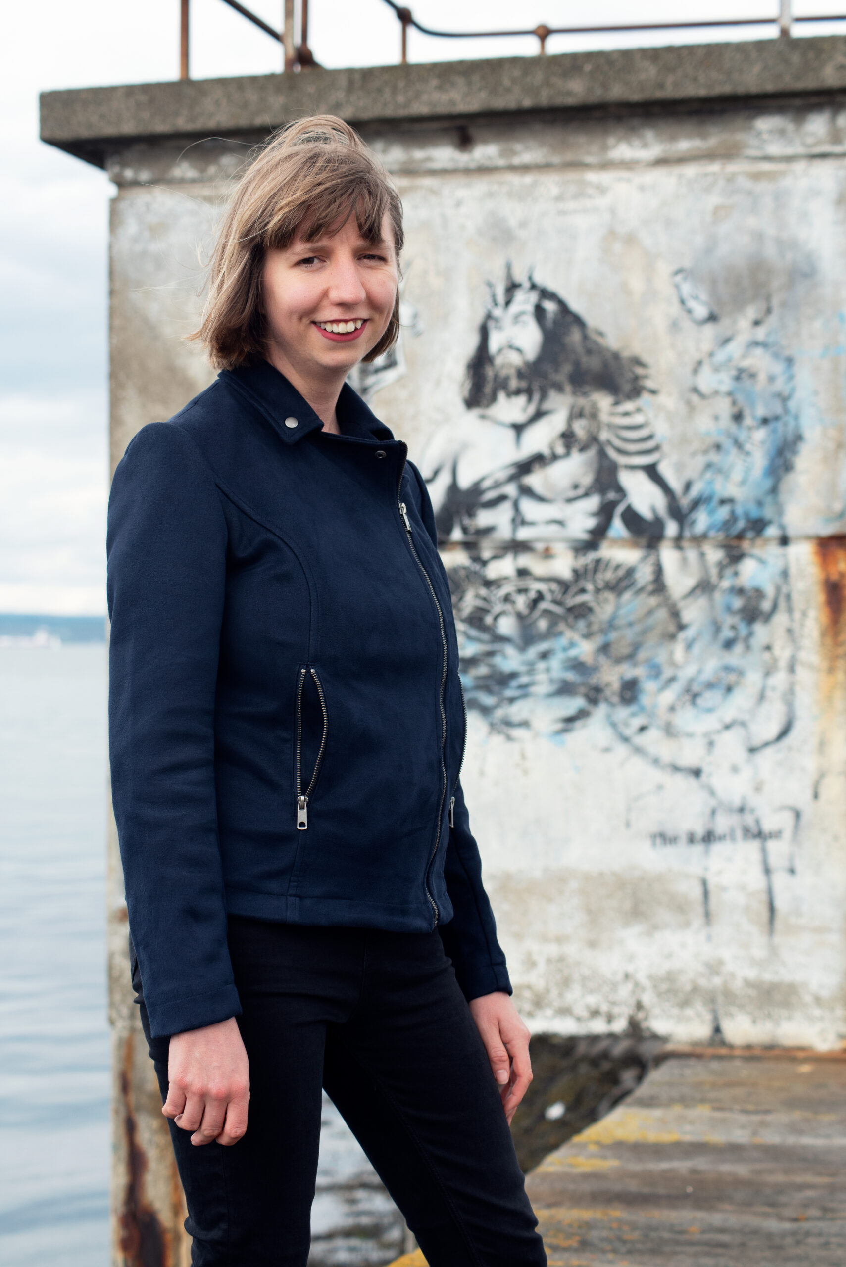 Translator Annie Rutherford smiling in dark clothes in front of a sea wall with a Poseidon graffito