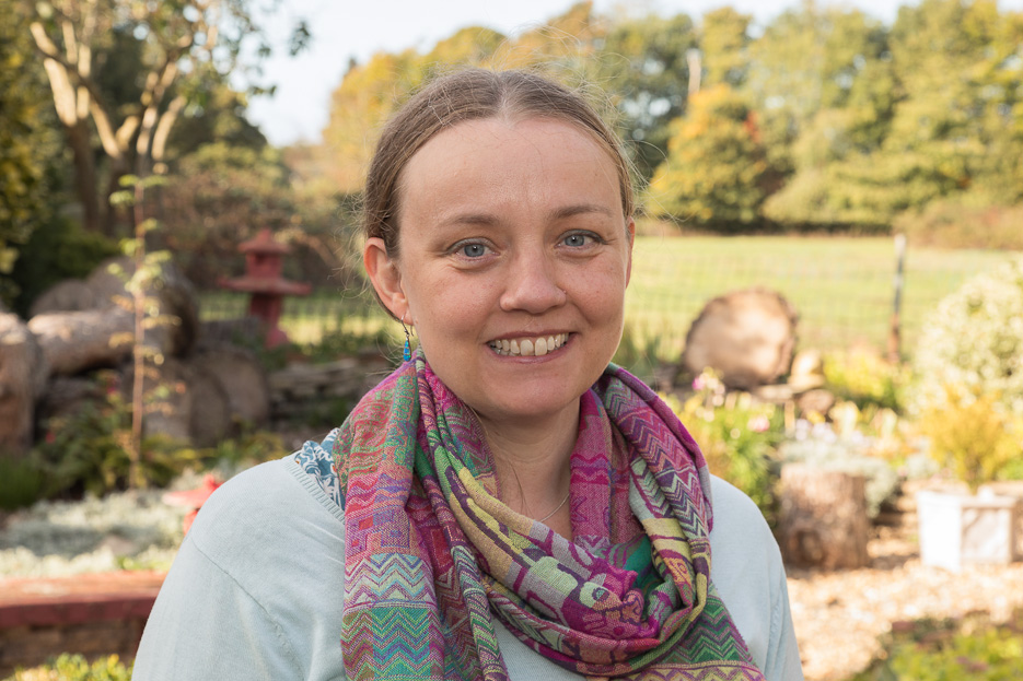 The translator Rachel Ward, a white woman with a brightly coloured summer scarf standing in front of a beautiful sunny garden, with a bird table in the background.