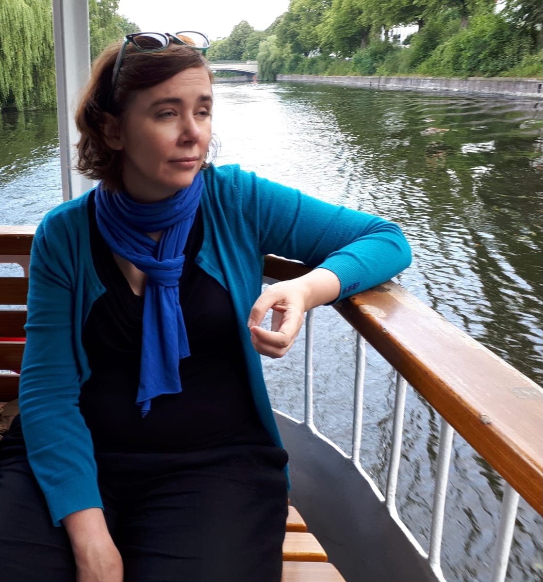 Translator Siobhán Dowling, a white woman with brown hair and a blue top, on board a boat on a river. She's wearing sunglasses on top of her head and looking melancholy.